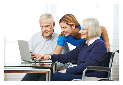 Female Nurse is standing between an elderly couple and they are all looking at an open laptop that is in front of them on a table